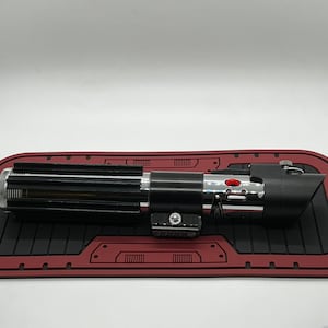 May include: A black lightsaber with a silver handle and red button. The lightsaber is on a red and black mat.