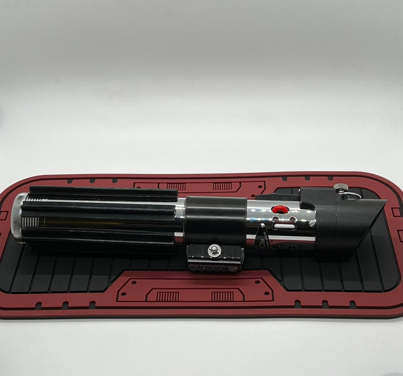 May include: A black lightsaber with a silver handle and red button. The lightsaber is on a red and black mat.