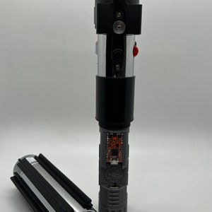 May include: A disassembled lightsaber with a black handle, silver blade, and a circuit board visible inside. The lightsaber is in two pieces, with the handle and blade separated.