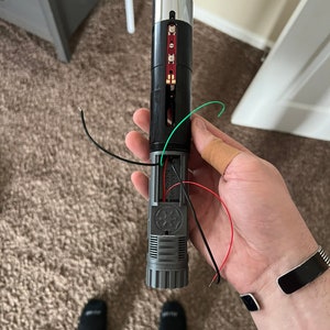 May include: A disassembled lightsaber with a silver tip, black body, and a red and green wire. The lightsaber has a black and gray handle with a Star Wars logo.