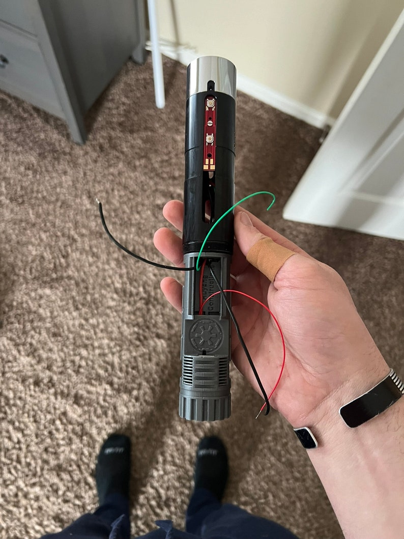 May include: A disassembled lightsaber with a silver tip, black body, and a red and green wire. The lightsaber has a black and gray handle with a Star Wars logo.