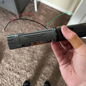 May include: A gray and black lightsaber with red, green, and black wires attached. The lightsaber is held in a hand with a bandage on the index finger.