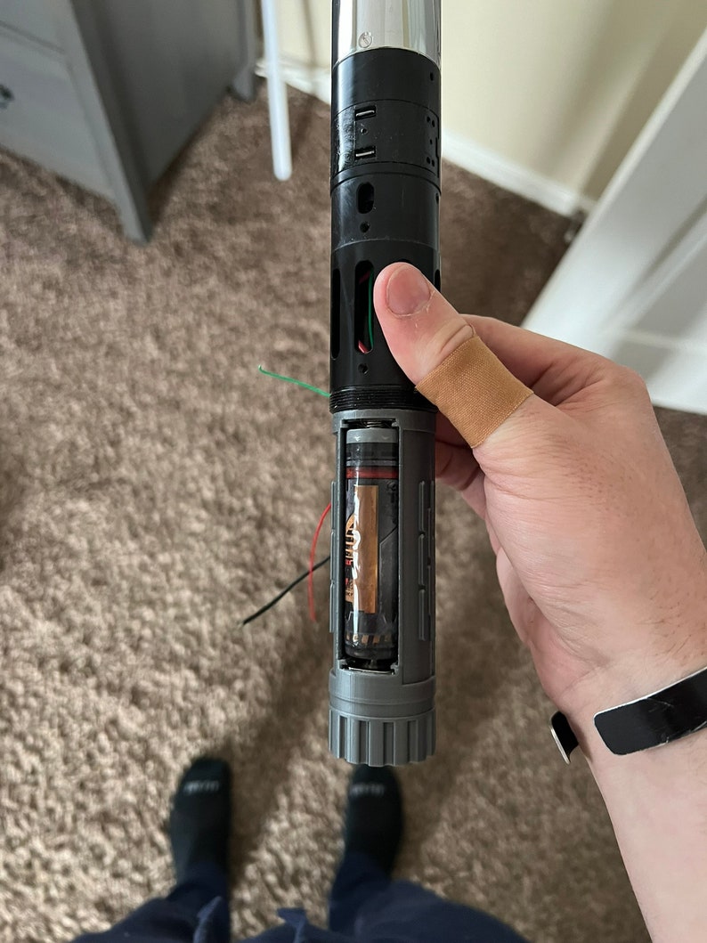 May include: A disassembled lightsaber with a silver and black handle. The handle is open, revealing a battery and wires. The lightsaber is held in a hand with a bandage on the thumb.
