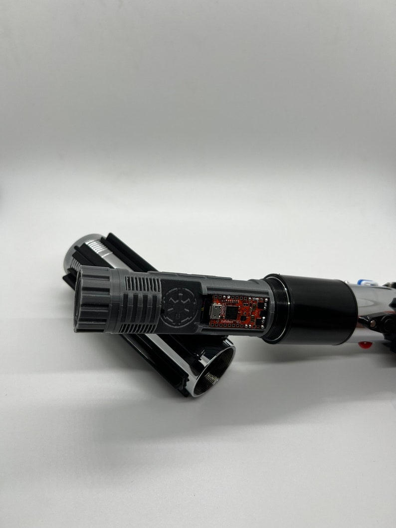 May include: A disassembled lightsaber with a black and silver hilt, a gray soundboard with a Star Wars logo, and a circuit board visible inside the hilt.