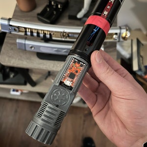 May include: A disassembled lightsaber with a gray and black hilt, a red and black lightsaber blade, and a circuit board with the Star Wars logo. The lightsaber is held in a person's hand.