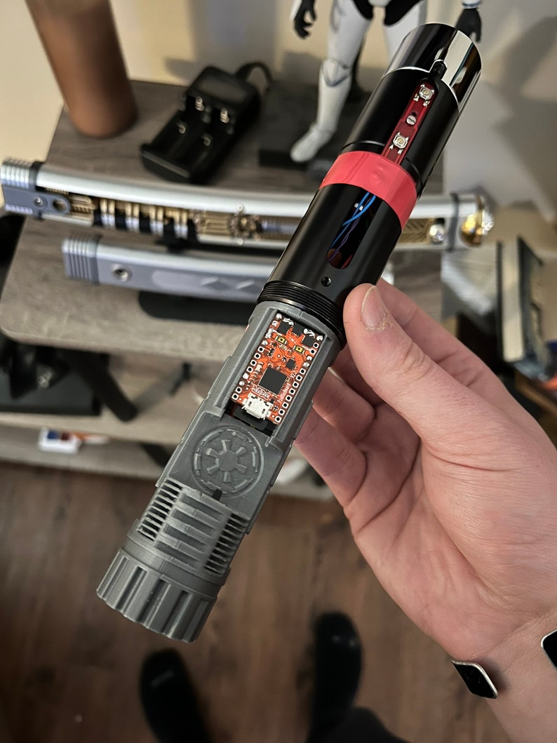 May include: A disassembled lightsaber with a gray and black hilt, a red and black lightsaber blade, and a circuit board with the Star Wars logo. The lightsaber is held in a person's hand.