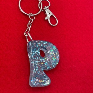 May include: A blue letter "P" keychain with iridescent glitter embedded in clear resin. The keychain has a silver-colored split ring and lobster clasp. The background is a solid red color.