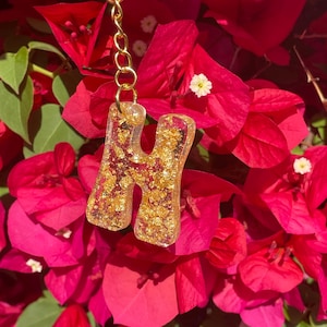 May include: A gold-colored letter "H" keychain with gold flakes suspended in clear resin. The keychain is attached to a gold chain and ring. The background features bright pink bougainvillea flowers.