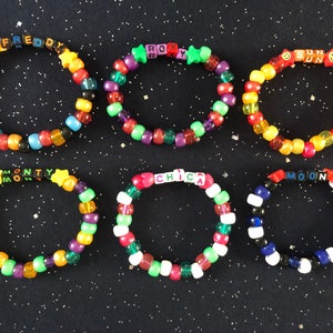 May include: Six colorful beaded bracelets with different names spelled out on them. The names are Freddy, Roxy, Sun, Monty, Chica, and Moon. The bracelets are on a black background with glitter.