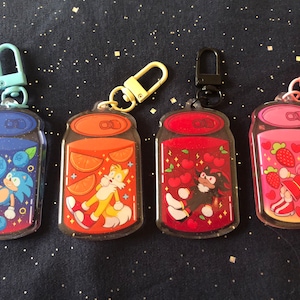 May include: Four colorful keychains featuring characters from the Sonic the Hedgehog video game series. Each keychain is shaped like a can of soda with a different character and flavor. The keychains are blue, orange, red, and pink.