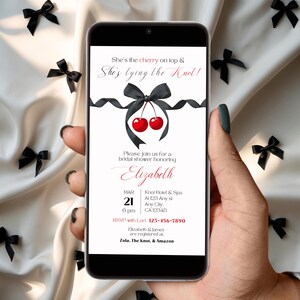 Op de afbeelding: Een digitale uitnodiging voor een bruidsdouche met een zwart lint en twee rode kersen erop. De tekst luidt "She's the cherry on top & She's tying the knot!" De uitnodiging is voor Elizabeth's bruidsdouche op 21 maart om 18:00 uur in het Knol Hotel & Spa. RSVP met Lori op 123-456-7890. Elizabeth & James zijn geregistreerd bij Zola, The Knot, & Amazon.