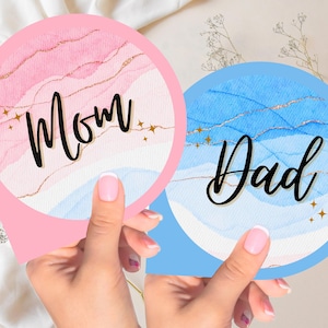 May include: Two round gender reveal signs with watercolor designs. The pink sign says "Mom" and the blue sign says "Dad".