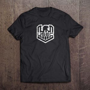 Puede incluir: Camiseta negra con un gráfico blanco. El gráfico presenta un diseño de águila con el texto "USEC" en forma de escudo. La camiseta está hecha de un material suave y se muestra sobre una superficie de madera.
