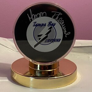May include: A black hockey puck with a gold and blue Tampa Bay Lightning logo and the signature "Martin St. Louis" in black ink. The puck is displayed in a clear plastic case on a gold metal stand.