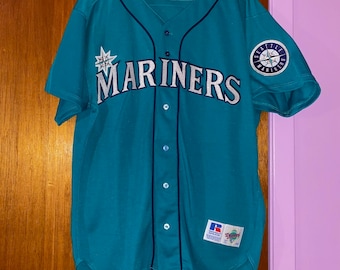 Może przedstawiać: Turkusowa koszulka baseballowa z napisem "MARINERS" w białym skrypcie na piersi. Koszulka ma zapinany na guziki przód, granatowe wykończenia i naszywkę na rękawie z napisem "SEATTLE MARINERS". Widoczna jest metka Russell Athletic.