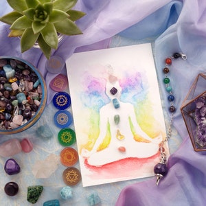 May include: A watercolor painting of a person in a yoga pose with seven chakra symbols and stones. The painting is surrounded by crystals, a pendulum, and a small glass box with amethyst crystals.