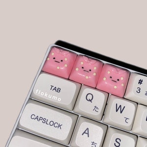 May include: A white keyboard with pink keycaps featuring cartoon faces. The keycaps are labeled with the letters Q, W, E, A, S, D, Z, and the words "TAB", "CAPSLOCK", and "SHIFT".