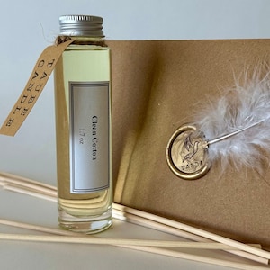 May include: A clear glass bottle of Clean Cotton oil with a silver cap and a brown tag labeled TAUBE CANDLE. The bottle is 1.7 oz. Beside it is a brown envelope with a gold wax seal featuring a bird and the word TAUBE, plus a white feather.