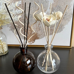 May include: Two round glass diffuser bottles with reed sticks. The left bottle is dark brown with straight and wavy black and beige reeds. The right bottle is clear with straight reeds topped with cream-colored wooden flower-shaped diffusers.