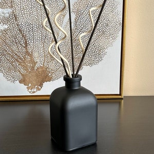 May include: Black diffuser bottle with a square base and a set of black and white diffuser sticks. The bottle is sitting on a dark wood surface.