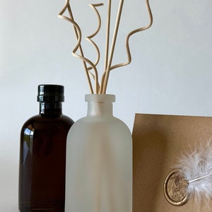 May include: A frosted glass diffuser bottle with a brown glass bottle in the background. Both bottles have black lids. The diffuser bottle has natural wooden sticks sticking out of the top. The sticks are curled at the top.