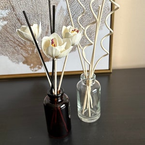 May include: Two glass bottles with diffuser sticks. One bottle is brown glass with black sticks and white flower accents. The other bottle is clear glass with natural colored sticks.