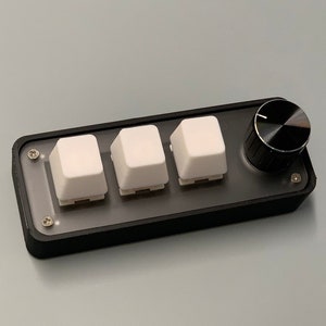 May include: A black control panel featuring three white square buttons and a black rotary knob. The panel has a clear plastic cover and is secured with small screws. The knob and buttons are on a black rectangular base.