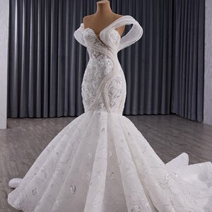 May include: A white, off-the-shoulder wedding gown with a mermaid silhouette. The bodice is embellished with intricate beadwork and floral appliques. The skirt flares out into a dramatic train, creating a romantic and elegant look.