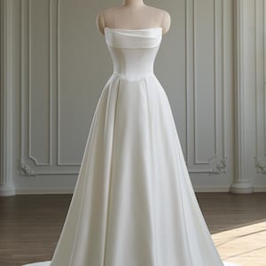 May include: A strapless white satin wedding dress displayed on a mannequin. The dress features a straight neckline with a folded-over detail, a fitted bodice, and a full A-line skirt that flows to the floor. The dress has a simple, elegant design, and is displayed in a well-lit room.