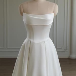 May include: A strapless white satin wedding dress displayed on a mannequin. The bodice features a folded, draped design across the bust and a fitted waist. The skirt flows into a full, A-line shape. The dress is simple and elegant, suitable for a bridal gown.