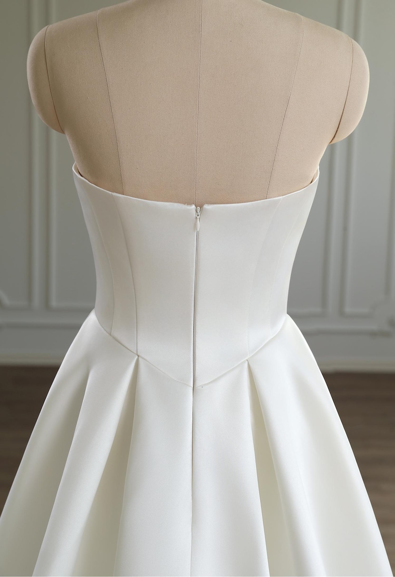 May include: A strapless, white wedding dress bodice on a mannequin. The bodice has a fitted, corset-like shape with a slight curve at the waist. The back features a zipper closure. The skirt of the dress begins to flare out from the waistline, with pleats adding fullness.