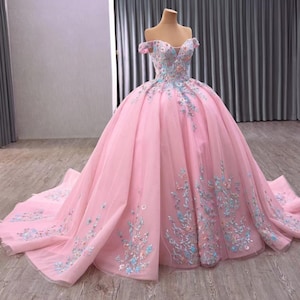 Può includere: Un abito da ballo rosa con spalle scoperte e ricami floreali. L'abito presenta uno scollo a cuore e una gonna ampia con strascico. I ricami includono disegni floreali blu e bianchi.