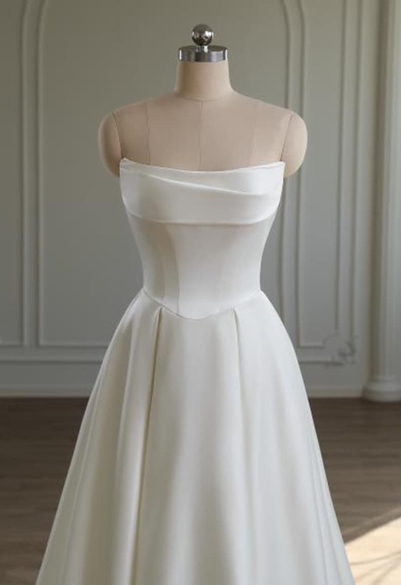 May include: A strapless white satin wedding dress displayed on a mannequin. The bodice features a folded, draped design across the bust and a fitted waist. The skirt flows into a full, A-line shape. The dress is simple and elegant, suitable for a bridal gown.