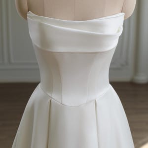 May include: A strapless white wedding dress displayed on a mannequin. The bodice features a structured, corset-like design with a curved neckline and a fitted waist. The skirt flares out from the waist, creating a classic A-line silhouette. The fabric appears to be a smooth, satin-like material.