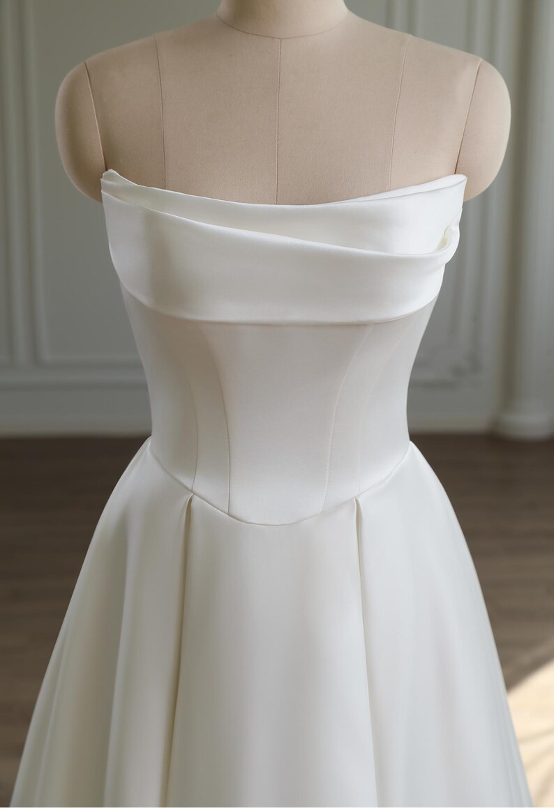 May include: A strapless white wedding dress displayed on a mannequin. The bodice features a structured, corset-like design with a curved neckline and a fitted waist. The skirt flares out from the waist, creating a classic A-line silhouette. The fabric appears to be a smooth, satin-like material.