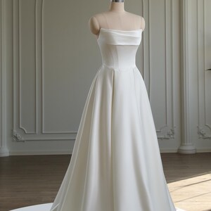 May include: A strapless, white satin wedding dress displayed on a mannequin. The dress features a straight neckline with a folded-over design and a fitted bodice. The skirt flows into a full A-line shape with a long train. The dress is in a studio setting with architectural details.