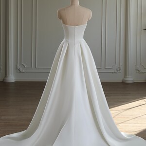 May include: A strapless white satin wedding dress displayed on a mannequin. The dress features a fitted bodice and a full, flowing skirt that extends into a long train. The back of the dress has a subtle V-shape and a zipper closure. The dress is set against a neutral background.