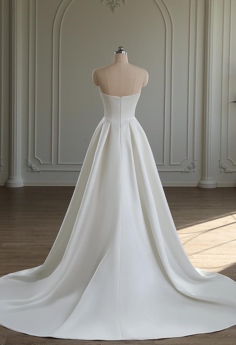 May include: A strapless white satin wedding dress displayed on a mannequin. The dress features a fitted bodice and a full, flowing skirt that extends into a long train. The back of the dress has a subtle V-shape and a zipper closure. The dress is set against a neutral background.