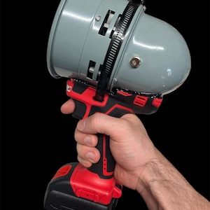 May include: A gray megaphone attached to a red and black power drill. The megaphone has a rounded top and a flared opening. The drill has a battery pack attached to the bottom. The image is set against a black background.