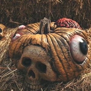 May include: A Halloween-themed pumpkin sculpture with a skull face, an eyeball, and a brain. The pumpkin is brown and sits on a bed of hay, with a skull in the background.