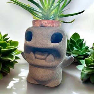 May include: A gray, textured, 3D printed planter shaped like a friendly ghost with a wide mouth and large eyes. A small green plant is growing in the planter.