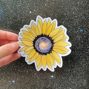 May include: A yellow sunflower with a galaxy swirling in the center. The sunflower is on a black background with white glitter.