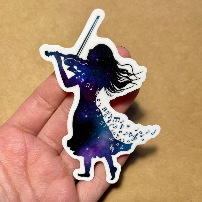 K&ouml;nnte beinhalten: Ein Vinyl-Aufkleber mit der Silhouette einer Person, die Geige spielt. Die Person ist von einer Galaxie aus Sternen und Noten umgeben.