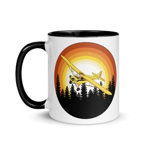 Puede incluir: Una taza de cerámica blanca con borde y asa negros. La taza presenta un diseño de puesta de sol retro con un avión amarillo volando sobre una silueta de árboles.