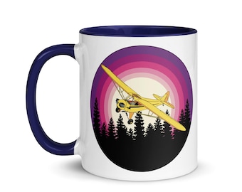 May include: A white ceramic mug with a blue rim features a yellow airplane flying over a forest silhouette against a sunset background with pink and purple hues.