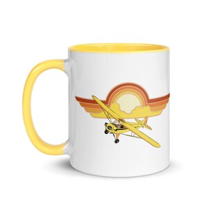 Könnte beinhalten: Eine weiße Keramiktasse mit gelbem Rand und Henkel. Die Tasse zeigt eine Retro-Illustration eines gelben Flugzeugs, das vor einem Sonnenuntergang mit orangefarbenen, roten und gelben Wolken fliegt.