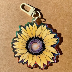 Może przedstawiać: Żółty słonecznik z galaktyką wirującą w środku. Słonecznik znajduje się na przezroczystym, tęczowym breloczku.