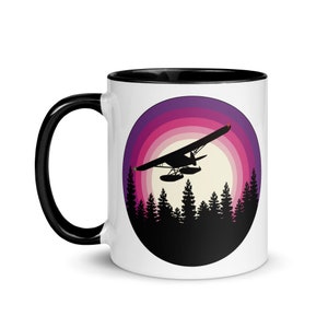以下が含まれることがあります： 黒色の縁取りと取っ手のある白いセラミックマグカップ。マグカップには、ピンクとパープルの夕焼けを背景に、森の上を飛ぶ飛行機のグラフィックが描かれています。