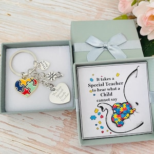 May include: A gift box with a silver keychain inside. The keychain has a heart-shaped charm with a puzzle piece design, a flower charm, and a heart charm with the text "The Best Special Needs Teacher". The box also contains a card with the text "It takes a Special Teacher to hear what a Child cannot say".