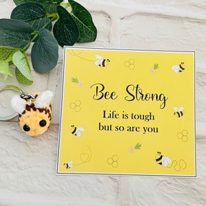 May include: A yellow card with the text "Bee Strong. Life is tough but so are you."  The card is decorated with small bees and honeycomb patterns. A crocheted bee keychain is also pictured.